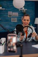 Smiling influencer recording podcast with headphones and microphone. Vlog headset review in front of recording smartphone. Content creator in home studio filming for social media. photo