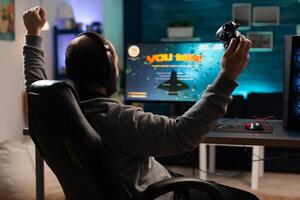 Person playing games with controller and winning in front of computer. Gamer using joystick and headphones to play online games. Man wearing headset and celebrating win. photo
