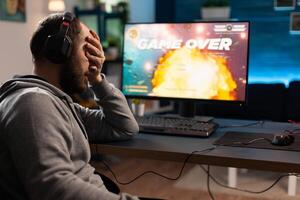 Sad person losing online games on computer. Man playing games with headphones and controller, feeling disappointed about lost game. Player using joystick and headset at monitor. photo