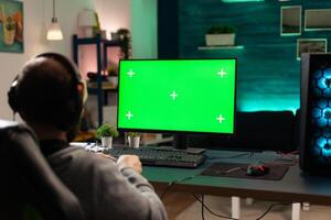 Player looking at computer with horizontal green screen. Man using isolated template and mockup background with chroma key to play games on monitor. Gamer holding controller. photo
