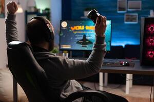 Man winning games with joystick and headphones in front of monitor. Gamer using controller and headset, playing online games on computer. Person celebrating game win for leisure. photo