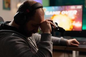 Sad gamer losing games after playing with controller and headphones on computer. Man feeling disappointed about lost game, using controller and headset. Player gaming with equipment photo