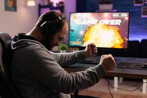 Disappointed man losing games on computer. Adult clenching fists and playing games with headphones and joystick, feeling sad about lost game. Player using modern equipment. photo
