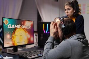 Couple of gamers feeling sad about losing games in front of computer. Woman comforting man about lost game online, having gaming equipment and headphones to play for leisure. photo