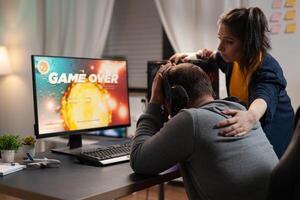 Couple feeling sad about losing games play in front of computer. Woman comforting man about lost game online, having gaming equipment and headphones, playing for leisure. photo