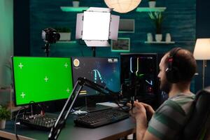 Man streaming games and using horizontal green screen. Person playing online games while he has chroma key with isolated mockup template on computer display. Gamer on stream photo