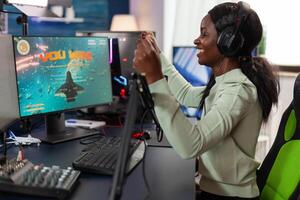 Happy african american woman gamer wearing headset winning videogames competition talking into microphone with players. Excited plater playing space shooter games using RGB computer. Live streaming photo