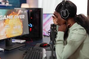 Sad upset woman gamer playing space shooter videogames losing online tournament while talking with players on streaming chat. Nervous streamer using RGB gaming computer. Game over on screen photo