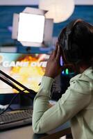 African american woman gamer losing online videogames during space shooter championship. Pro streamer wearing headphones talking with players on live chat playing game using RBG computer. Esport games photo