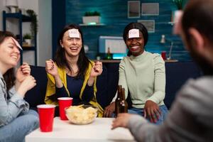 Laughing group of friends playing gusess game using stickey notes enjoying spending time together during wekeend party at home. Multi-ethnic people drinking beer guessing correct answer photo
