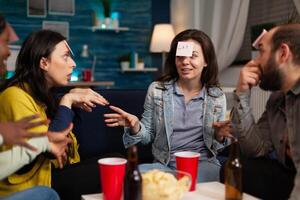 Portrait of happy woman with stickey notes on forehead playing guess who game enjoying spending time with friends socializing together during wekeend party at home. Friendship concept photo