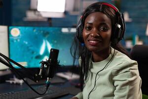 Portrait of african american player smiling at camera while playing space shooter videogame during online championship. Cyber player talking at microphone for streaming live using RGB computer photo