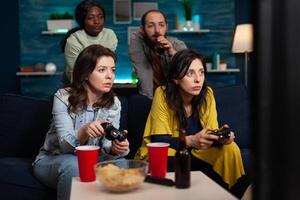Concentrated women relaxing on couch playing games using controller during online competition enjoying spending time together in living room. Group of multi-ethnic friends having fun at home photo