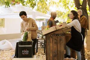 Courier putting package of organic fruits and vegetables delivery. in thermal food bag while standing near market counter, picking up order. Farmers deliver fresh produce to local customers photo