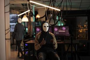 Cybercriminal in mask hacking computer password to steal information, breaking into online server. Anonymous hacker attacking database and committing cyberattack late at night. photo