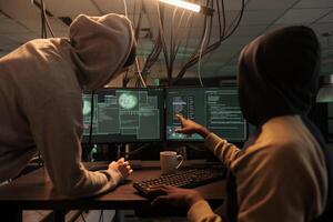 Internet criminals hacking online server, stealing finances, cyberattack. Hacker in hoodie pointing at code on pc screen, scammers programming malicious software at night time photo