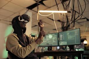African american hacker working at night with 3d vr glasses, trying to break in government server with virtual reality. Male criminal using interactive headset to steal sensitive official data. photo