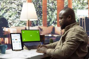 Business owner working at home with a chroma key display on laptop, taking notes after doing financial report and accounting analytics. Startup administrator using green screen at desk. photo