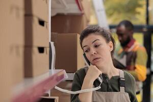 Storage room employee talking at landline phone with remote business owner before start working at clients packages shipment. Worker doing products quality control in warehouse. Delivery concept photo