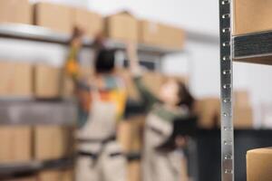 Selective focus of carton boxes standing on shelves, in background storehouse employees preparing customers orders. Storage room team managing parcels transportation, working in warehouse photo