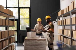 Diverse warehouse employee analyzing goods logistics on laptop computer, working at merchandise quality control report. Stockroom supervisors wearing protective overall and helmet preparing orders photo