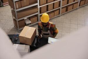 Top view of diverse employees discussing clients orders preparing packages for delivery in storehouse. African american worker wearing industrial overall and helmet working at goods quality control photo
