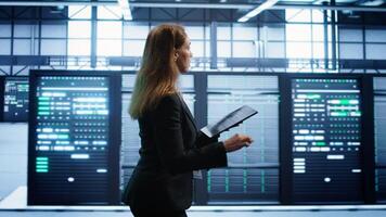 Engineer in server hub checking recovery plan on tablet, monitoring rackmounts energy consumption. Woman in data center making sure rigs are functioning optimally, doing routine disk checks, camera B photo