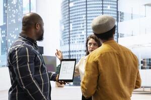 Real estate broker presenting a new high class office space to clients, discussing the detailed blueprints using a tablet during the office tour. Planning an investment strategy involving property. photo
