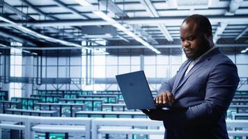 African american IT operator doing maintenance using laptop to prolong data center equipment life span. IT professional in server hub workspace monitoring server mainframes using notebook, camera B photo
