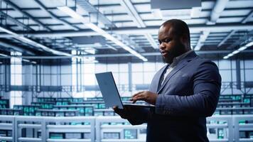 Programmer in data center typing on laptop, running error checking utilities. IT professional in server room looking for network issues affecting rigs performance using digital device, camera B photo