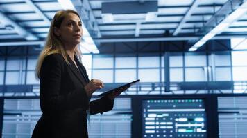 Engineer in server hub checking recovery plan on tablet, monitoring rackmounts energy consumption. Woman in data center making sure rigs are functioning optimally, doing routine disk checks, camera A photo