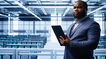 Programmer in server room location using tablet to patch network issues affecting transfer rates. African american man in data center fixing errors affecting mainframes, camera B photo