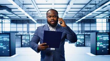 IT professional looking over data center checkup checklist paperwork, talking on phone in server farm. African american man surrounded by server rigs, chatting during telephone call, camera B photo
