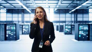 Software engineer talking on smartphone in data center, coordinating server maintenance tasks. Woman surrounded by server racks and high tech equipment, speaking during telephone call, camera B photo