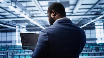 Programmer doing yearly maintenance checkup using laptop to prolong data center electronics life span. IT professional in server farm monitoring server infrastructure using notebook, camera B photo
