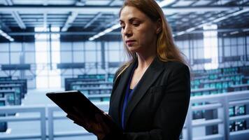 Admin in server room checks recovery plan on tablet, monitoring equipment energy consumption. IT professional uses notebook in data center workplace, ensuring rigs are functioning optimally, camera B photo