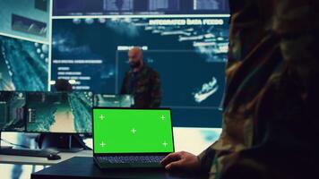Army officer studies isolated greenscreen display on a laptop in control room, military power base personnel monitoring satellite world map for federal operations. Commander in uniform. Camera B. photo