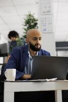 Arab project manager chating with coworker online on laptop while working in business office. Young financial analyst analyzing statistics report in corporate coworking space photo