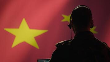 Chinese army military dispatcher looking at surveillance transmission on laptop, relaying informations for troops using headset, isolated over fluttering China flag background, camera B photo