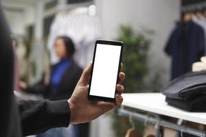 Shopping mall buyer hand holding smartphone with empty screen, browsing apparel online and in boutique. Clothing store customer using mobile phone with blank touchscreen mockup for promotion photo