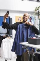 Clothing store smiling customer showing jacket on hanger while chatting in videocall using smartphone. Fashion blogger showing apparel while streaming online with mobile phone photo