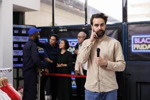 Young man TV reporter checking earphone looking at camera while making Black Friday news episode. Broadcast journalist reporting about crowd of shoppers waiting to get into store during seasonal sales photo