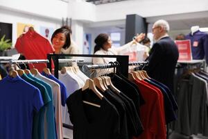 Seasonal sales. Clothes hanging on rack in clothing store with diverse people customers shopping at discounted prices on background. Retail fashion business and bargain hunting concept photo