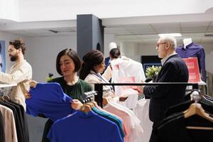 Customer service during Black Friday. Retail worker having conversation with senior man customer in crowded clothing store during seasonal sales, helping shopper to choose clothes at discounted prices photo