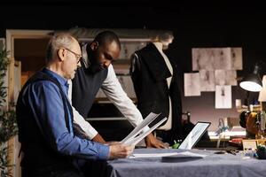 Tailors checking fashion sketch designs, trying different attire combinations on mannequin in haute couture atelier. Master craftsman and apprentice working on bespoke suit alterations photo
