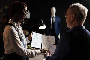 African american tailor apprentice and elderly master dressmaker comparing sketch drawing on tablet and paper, trying to design the perfect dress. Couturiers working on client comission photo