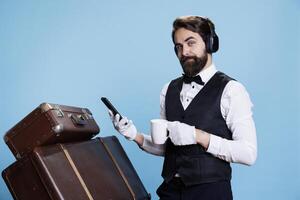 Hotel guard text and drinks coffee, checks emails on his mobile phone while he listens to tunes. Bellman employee navigating internet pages and sipping refreshing drink with headset on. photo