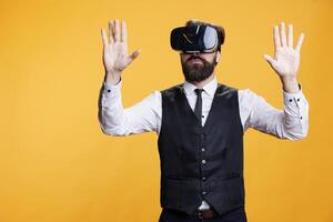 Modern waiter using virtual reality, enjoying 3d vr headset against yellow background. Young man employee having fun with interactive vision simulation on camera, innovative glasses. photo