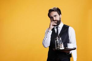Skilled waiter calculating bill in studio, preparing to serve clients with meal or refreshment against yellow background. Elegant restaurant butler acting thoughtful on camera. photo