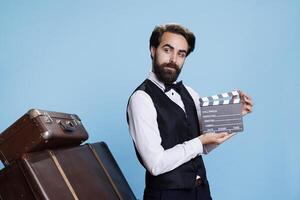 Confident doorman holds clapboard against blue background, clapping filmslate in studio for post production. Stylish bellboy using clapper board on camera, movie filming concept. photo